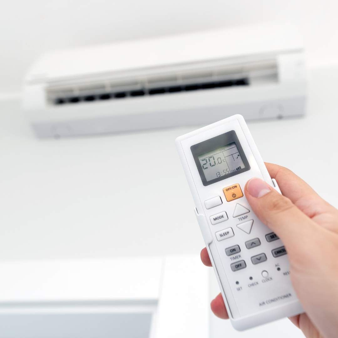 The blurred out image of a white, wall-mounted air conditioner with a hand in the foreground adjusting the temperature on a remote control.
